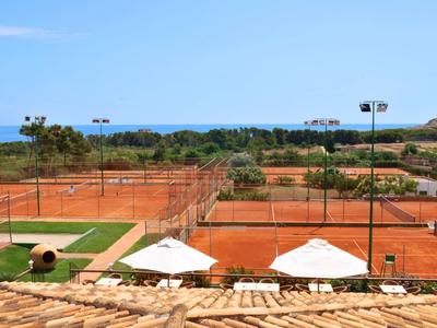 Multiple red clay tennis courts with two white umbrellas under clear sky.