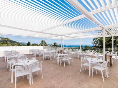 Open outdoor restaurant area with white tables and chairs under a white pergola overlooking the sea.