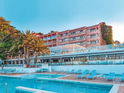 Modern hotel with outdoor pool, sun terrace, and palm trees under clear blue sky.