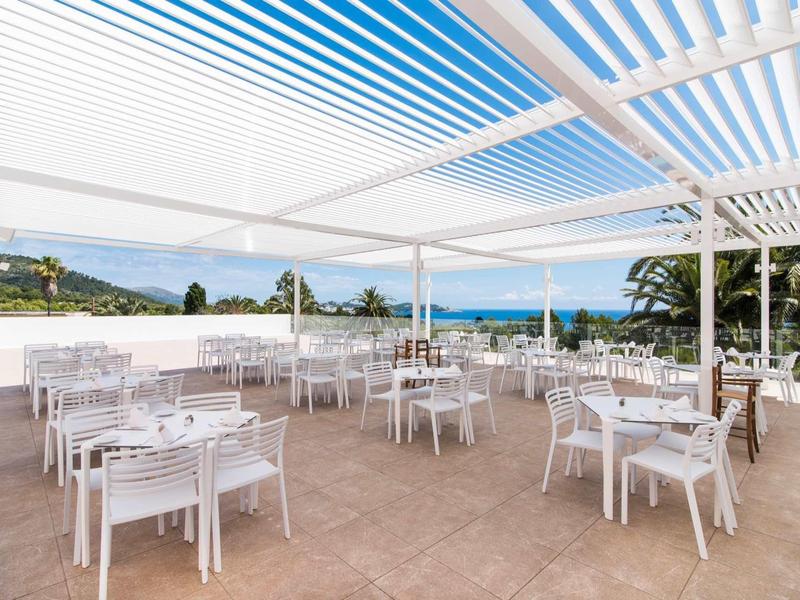 Open outdoor restaurant area with white tables and chairs under a white pergola overlooking the sea.