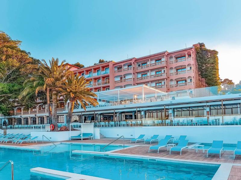 Modern hotel with outdoor pool, sun terrace, and palm trees under clear blue sky.