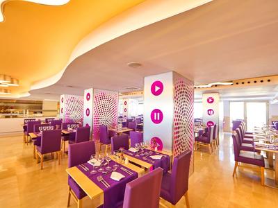 Heldere restaurantinterieur met paarse stoelen en gele tafelkleden onder moderne plafondlampen.