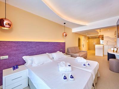 Lichte hotelkamer met tweepersoonsbed, paarse hoofdbord, moderne verlichting, fauteuil en kitchenette.