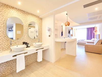 Ruime moderne hotelkamer met wastafels, zithoek en bed met paarse gordijnen.