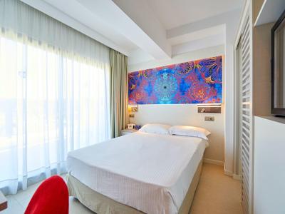 Lichte hotelkamer met een tweepersoonsbed, kleurrijke kunst en een groot raam met doorschijnende gordijnen.