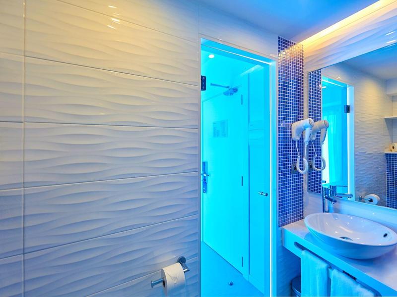 Moderne hotelbadkamer met blauw verlichte glazen douche, wastafel, spiegel en gestructureerde muren.