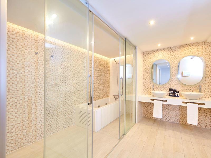 Moderne badkamer met glazen douche, dubbele wastafel en beige mozaïektegels aan de muren.