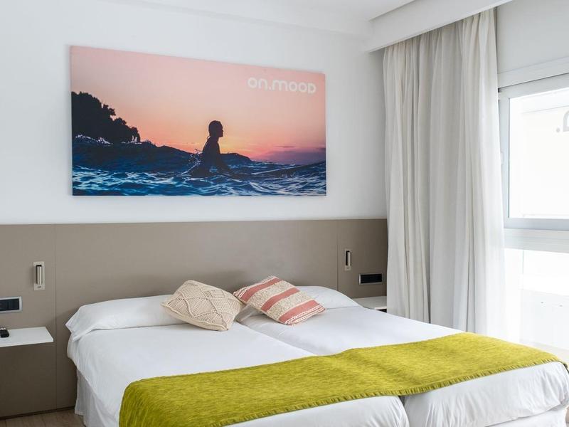 Jasny pokój hotelowy z podwójnym łóżkiem, zielonym narzutą i dużym oknem z białymi zasłonami.