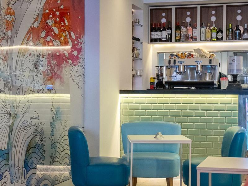 Przytulna strefa lounge hotelu z niebieskimi fotelami, stołami i barem w tle.