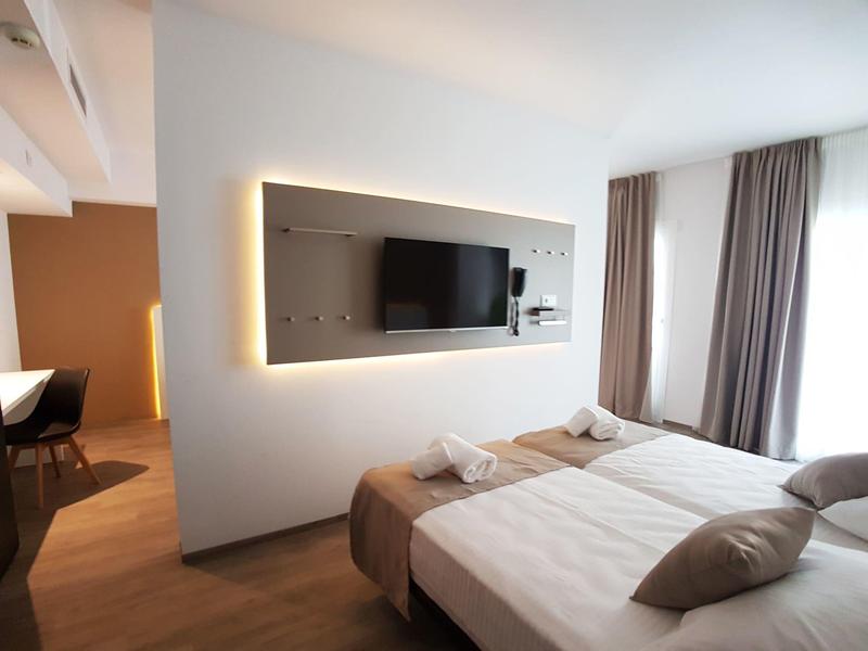 Camera d'hotel moderna con letto matrimoniale, TV e luce naturale da due finestre.