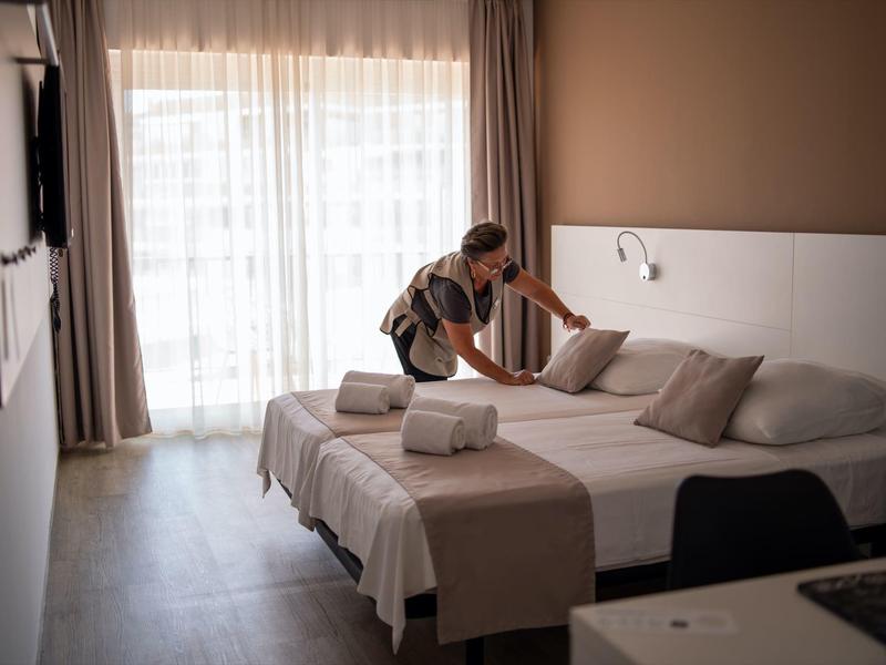 Persona che sistema un letto in una camera d'hotel moderna e luminosa con tende e colori caldi.