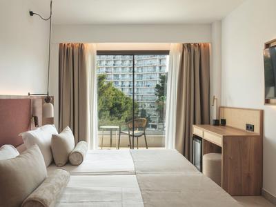 Hotel room with two beds, sofa, and balcony overlooking a garden and a building.