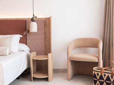 Modern hotel room area with bed, comfortable armchair, and bedside table in neutral tones.