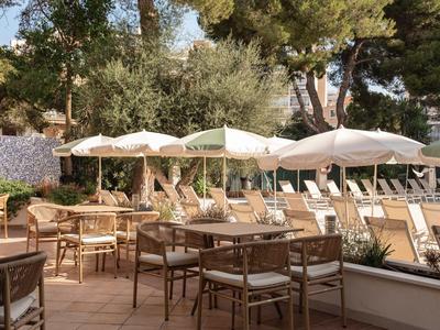Sunny terrace with tables, chairs, and umbrellas beside a pool and trees.