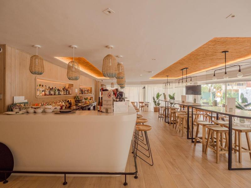 Světlý, moderní hotelový bar s dřevěným obložením, bílými pulty a vysokými stoličkami za denního světla.