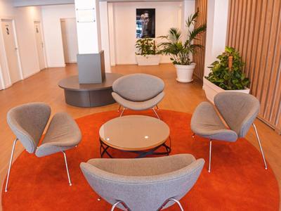 Nowoczesna poczekalnia z szarymi fotelami, okrągłym drewnianym stołem i pomarańczowym dywanem.