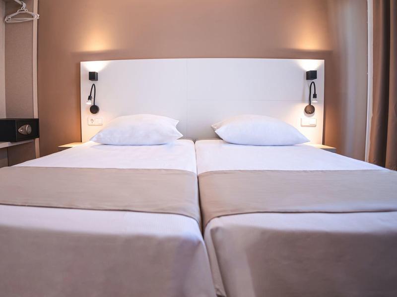 Dwa pojedyncze łóżka z białą pościelą i wspólnym zagłówkiem w nowoczesnym pokoju hotelowym.