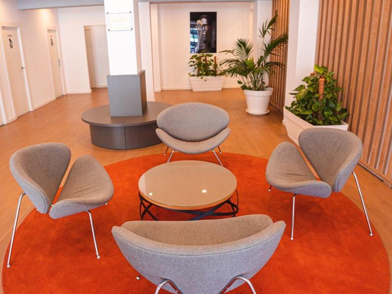 Nowoczesna poczekalnia z szarymi fotelami, okrągłym drewnianym stołem i pomarańczowym dywanem.