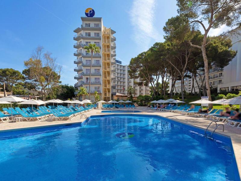 Hôtel avec grande piscine, entourée de chaises longues et de grands arbres sous un ciel bleu.