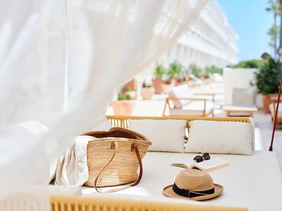 Luminoso balcone dell'hotel con cappello di paglia e borsa su divano bianco al sole.