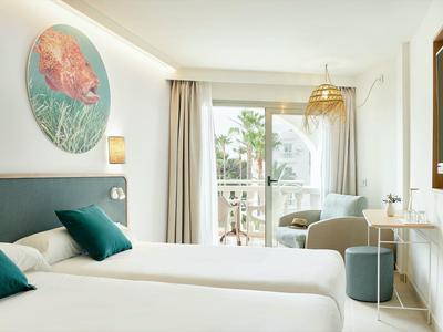 Camera d'hotel luminosa con due letti, cuscini verde-blu e balcone con vista sulle palme.