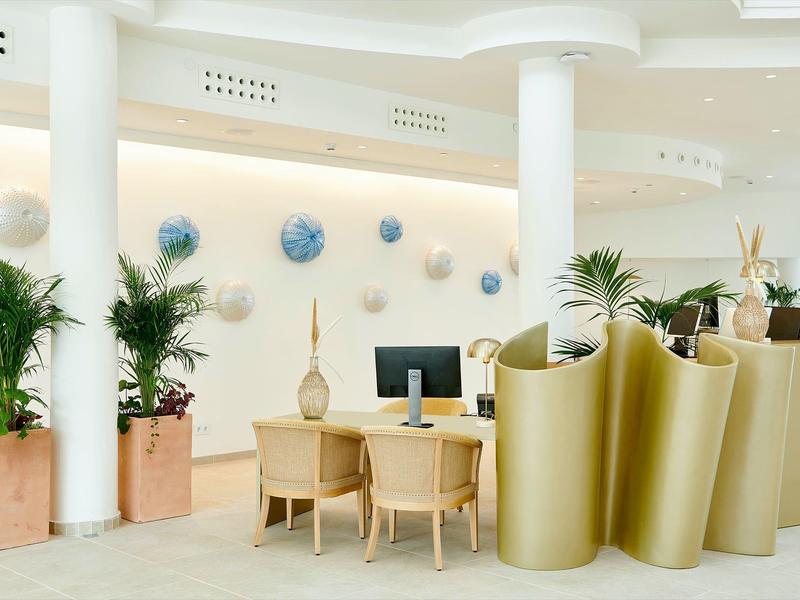 Lobby dell'hotel luminosa con aree salotto, piante e computer vicino a colonne bianche.