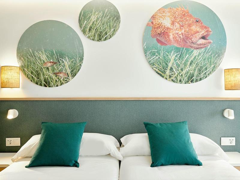 Camera d'albergo con due letti, cuscini verdi e quadri murali a tema marino.