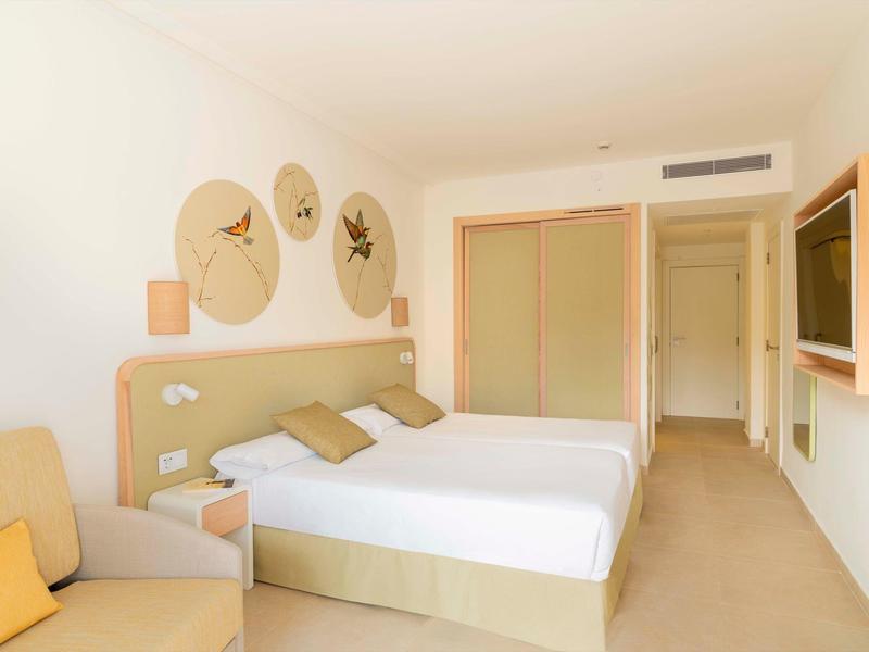 Camera d'albergo luminosa con due letti singoli, divano e armadio a muro in tonalità neutre.