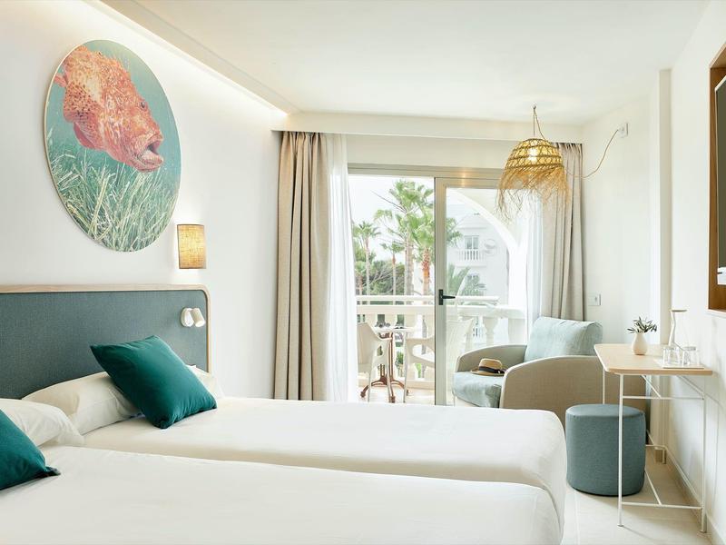 Camera d'hotel luminosa con due letti, cuscini verde-blu e balcone con vista sulle palme.