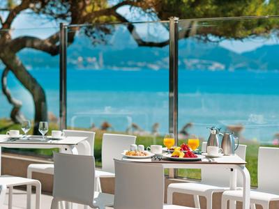 Restaurantterrasse mit modernen weißen Tischen und Stühlen, Meerblick und Bergen im Hintergrund.