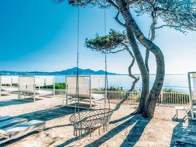 Schaukel am Baum mit Meerblick, blauer Himmel, weiße Liegestühle und Strandbars im Hintergrund.