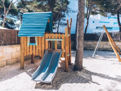 Kinderspielplatz mit Rutsche, Holzhaus und Schaukel im Sand unter Bäumen an sonnigem Tag.