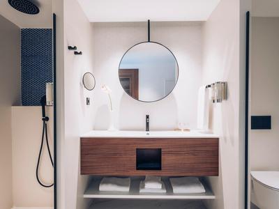 Moderner Badezimmerschrank mit rundem Spiegel, Holzschublade und Waschbecken, links Duschbereich sichtbar.