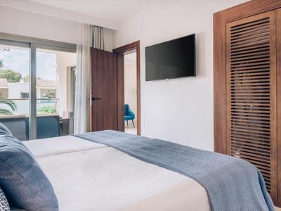 Modernes Schlafzimmer mit großem Bett, blauen Kissen, TV an der Wand und Balkon mit Gartenblick.