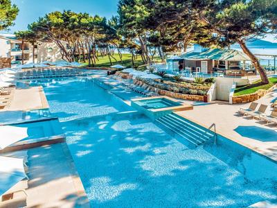 Großer blauer Pool mit weißen Liegestühlen, grünen Bäumen und Meerblick im Hintergrund.