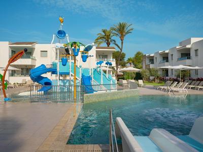 Pool mit Wasserrutschen, buntem Wasserspiel und Liegestühlen zwischen weißen Gebäuden.
