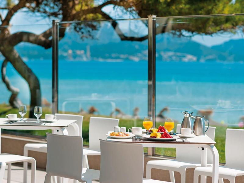 Mesa con desayuno en una terraza con vista al mar y árboles al fondo.