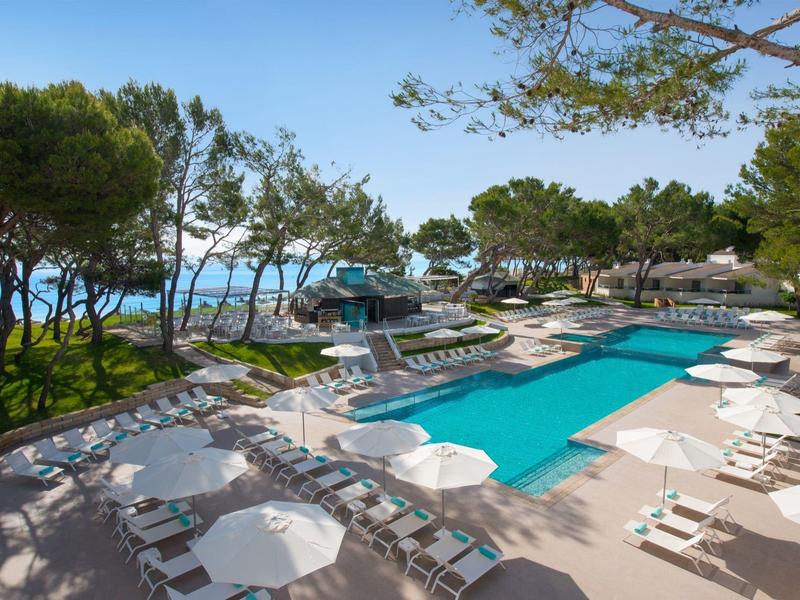 Una piscina moderna de hotel con tumbonas y sombrillas, rodeada de árboles con vista al mar.