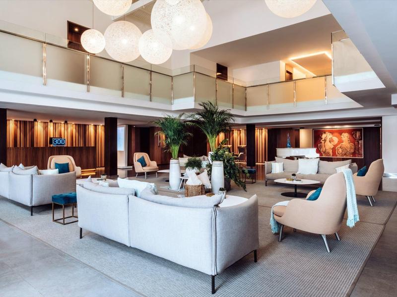 Amplia zona de salón de hotel con sofás blancos, sillones modernos y lámparas colgantes decorativas.