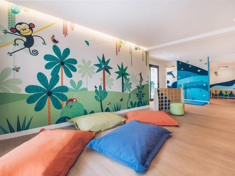 Sala infantil luminosa con cojines de colores y mural con flores y mono en un hotel.