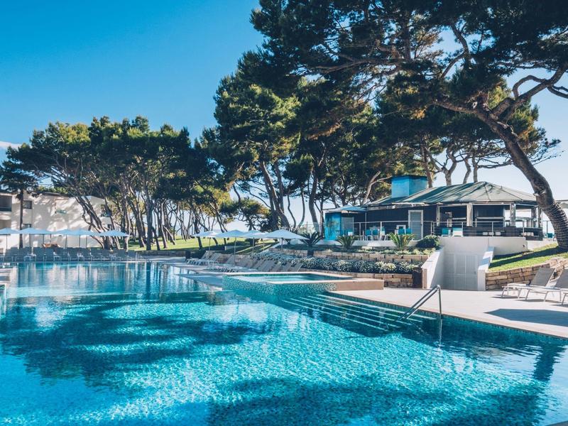 Piscina moderna de hotel con agua cristalina, tumbonas blancas y árboles al fondo bajo clima soleado.