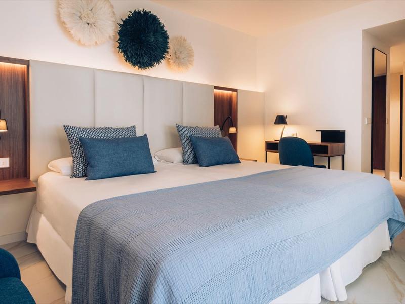 Habitación de hotel moderna con dos camas individuales, cojines azules y escritorio con luz cálida.