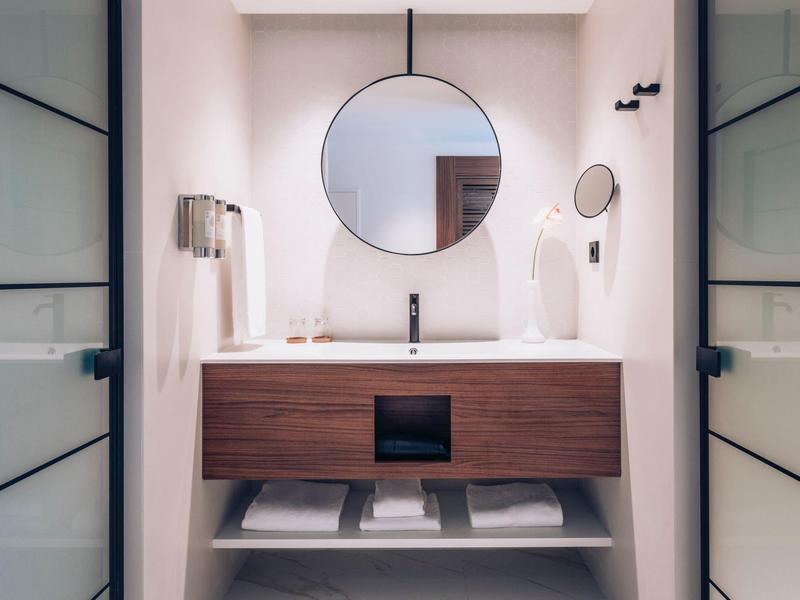 Baño moderno con espejo redondo, lavabo de madera y paredes blancas.