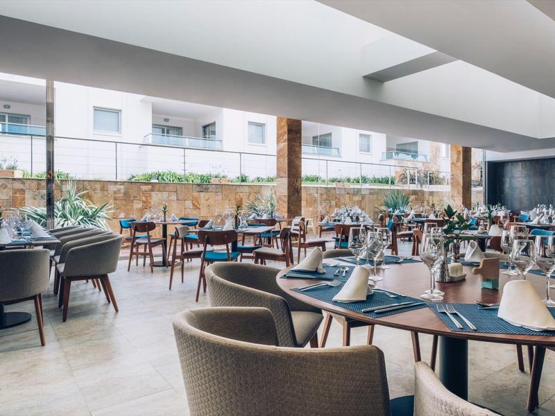 Restaurante de hotel moderno con grandes ventanales, mesas de madera clara y sillas acolchadas cómodas.