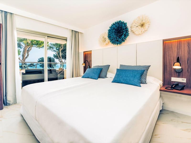 Habitación de hotel luminosa con cama grande, cojines azules y balcón con vista a árboles.