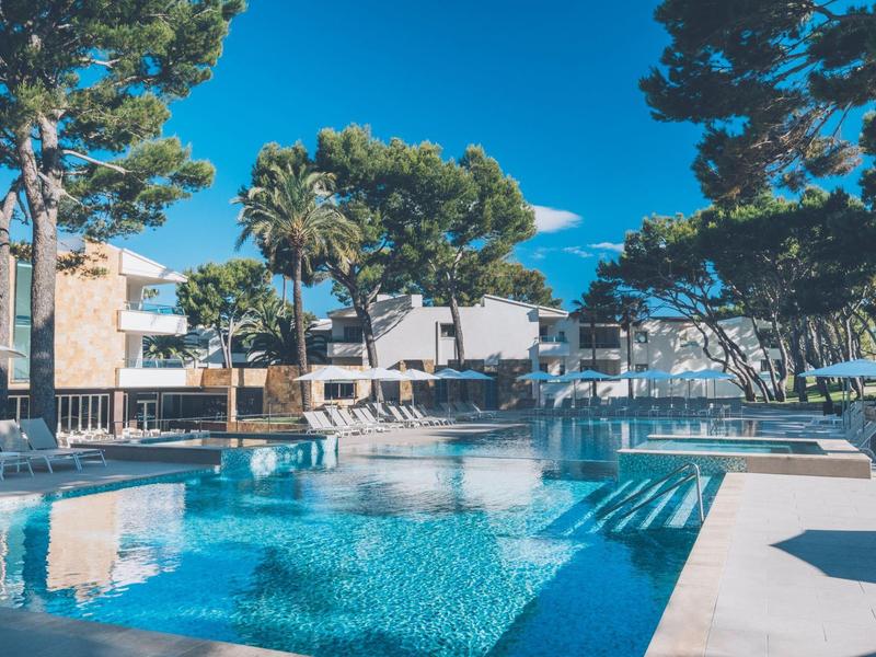 Piscina clara en un jardín de hotel con tumbonas y árboles bajo un cielo azul.
