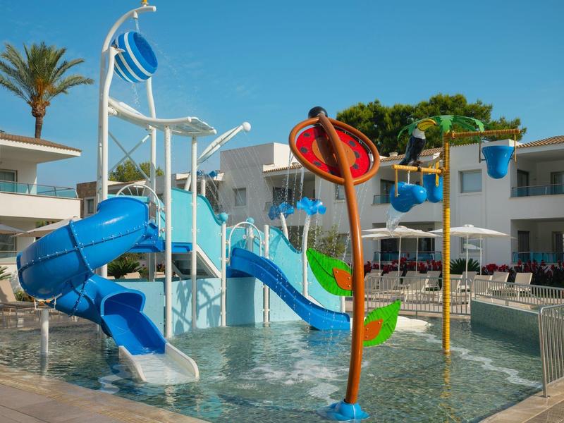 Área de juegos acuáticos para niños con toboganes y sombrillas de agua en entorno hotelero.