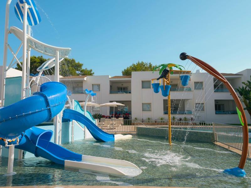 Área de piscina con dos toboganes de agua y juegos acuáticos frente a un edificio de hotel bajo cielo despejado