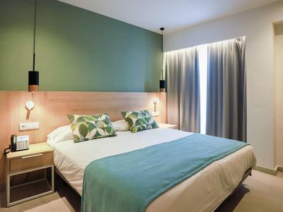 Habitación de hotel moderna con cama grande, pared verde y luz natural por ventana con cortinas.