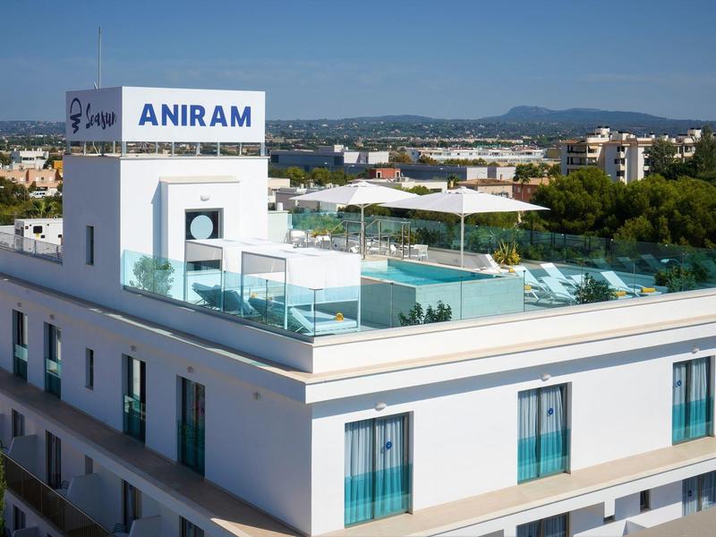 Hotel moderno con terraza en la azotea y piscina, rodeado de ciudad y montañas al fondo.